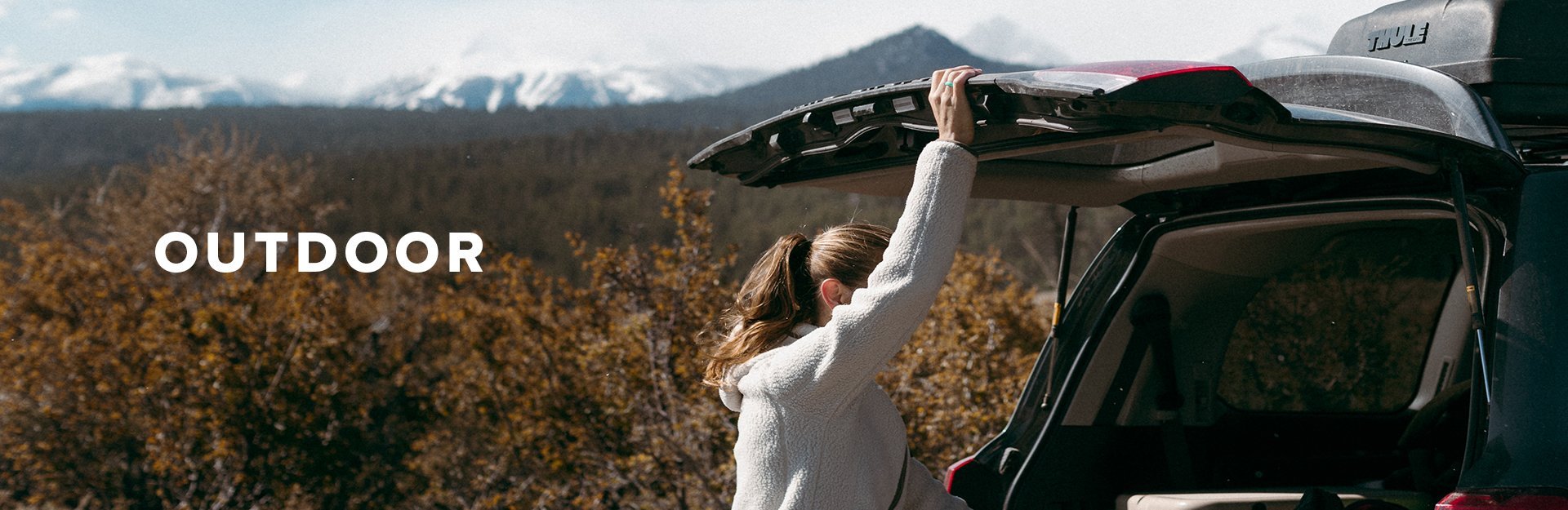 Outdoor, a woman holds up the hatch of her car with mountains in the background