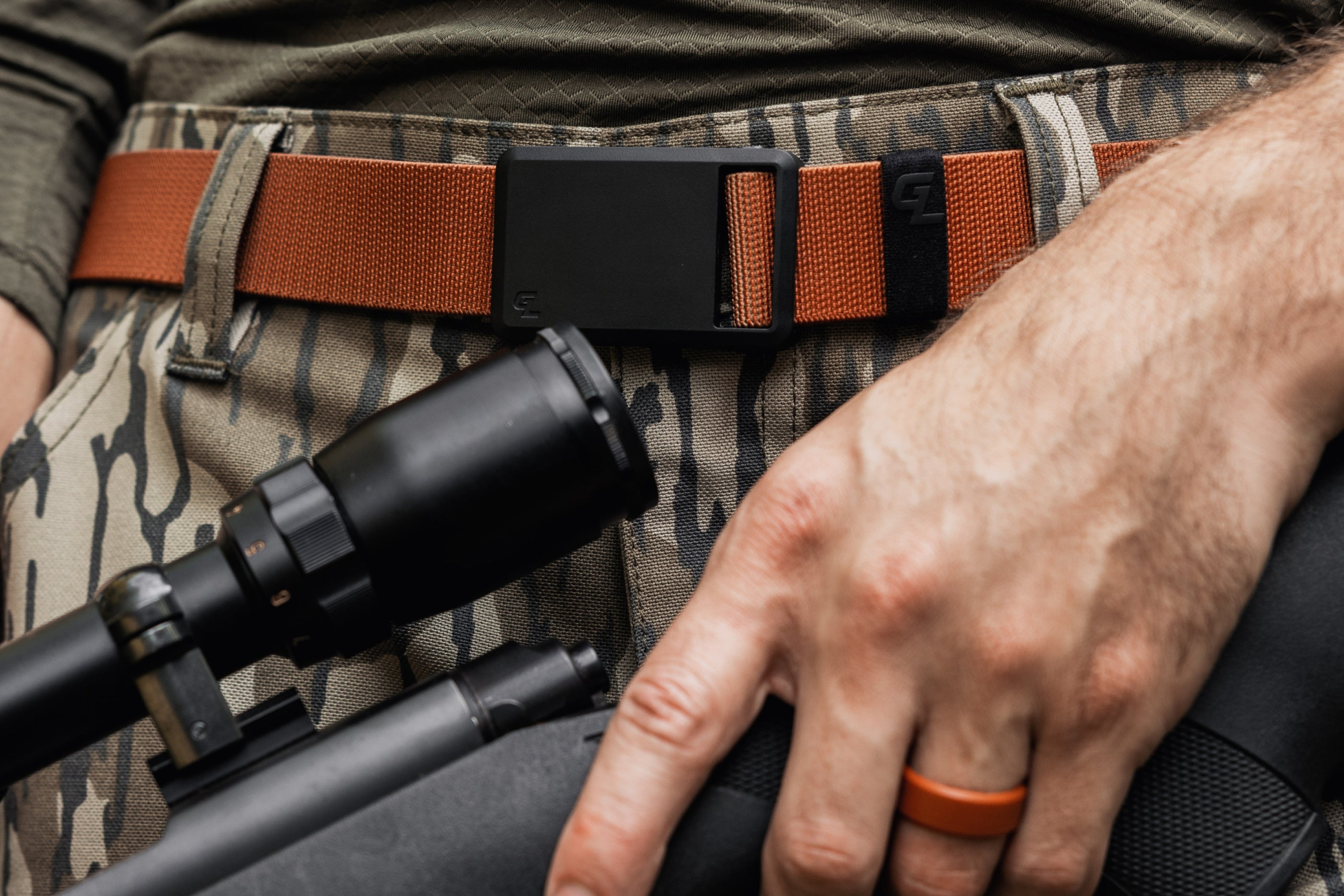 Person holding a rifle with a scope, wearing camouflage pants and an orange belt.
