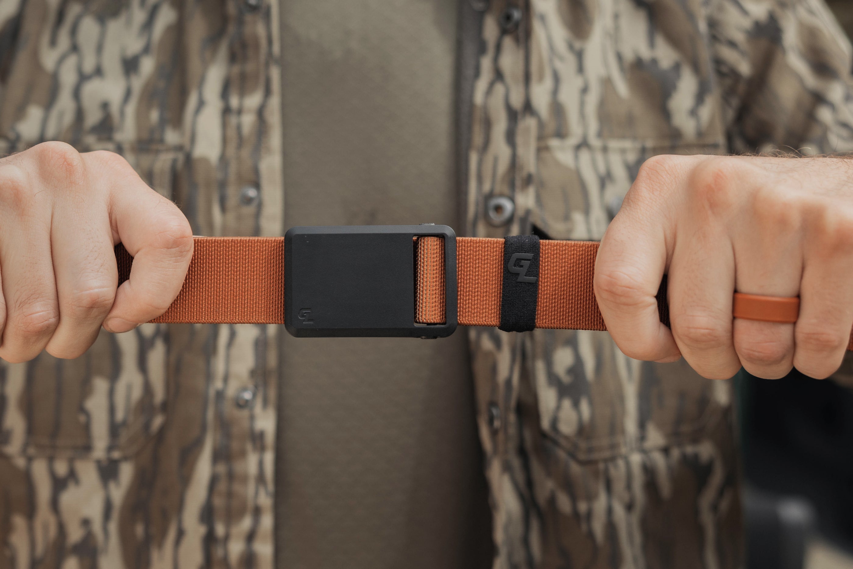Person holding a brown belt with a black buckle against a camouflage-patterned background
