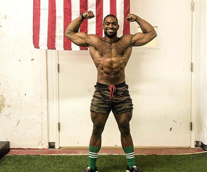 Andre Crews flexes his biceps in front of a white wall and american flag