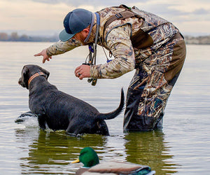 A man in camo points off camera for his duck dog, a black lab