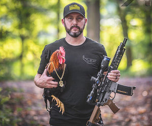 Justin Holt stands holding a large rifle in his left hand and a rooster wearing a gold chain in his right hand