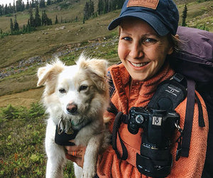 Caitlin Triana is sitting with her dog in a valley with a camera slung over her shoulder