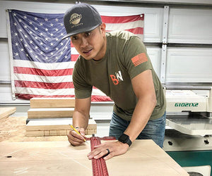 Justin Cunanan measures a block of wood in his woodshop, with an American flag in the background
